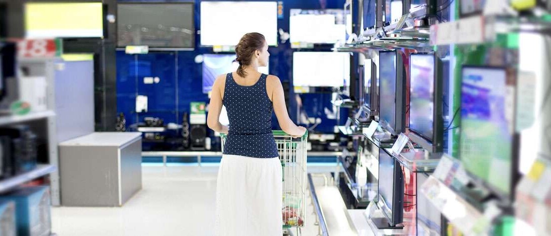 Woman shopping with cart in front of her as she browses a TV aisle at a durable goods retail store