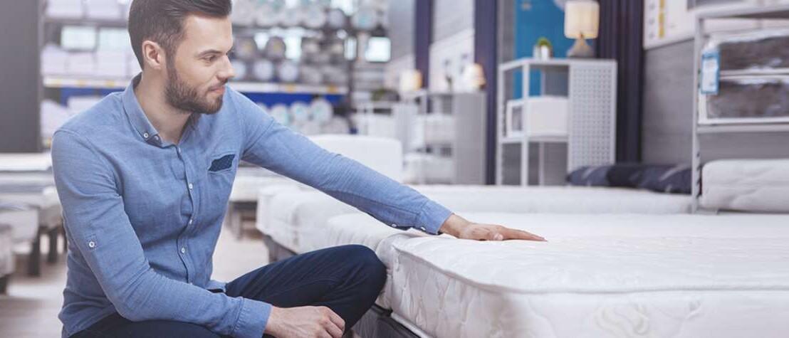 Man sits cross-legged by a mattress in a store with his hand on top of it